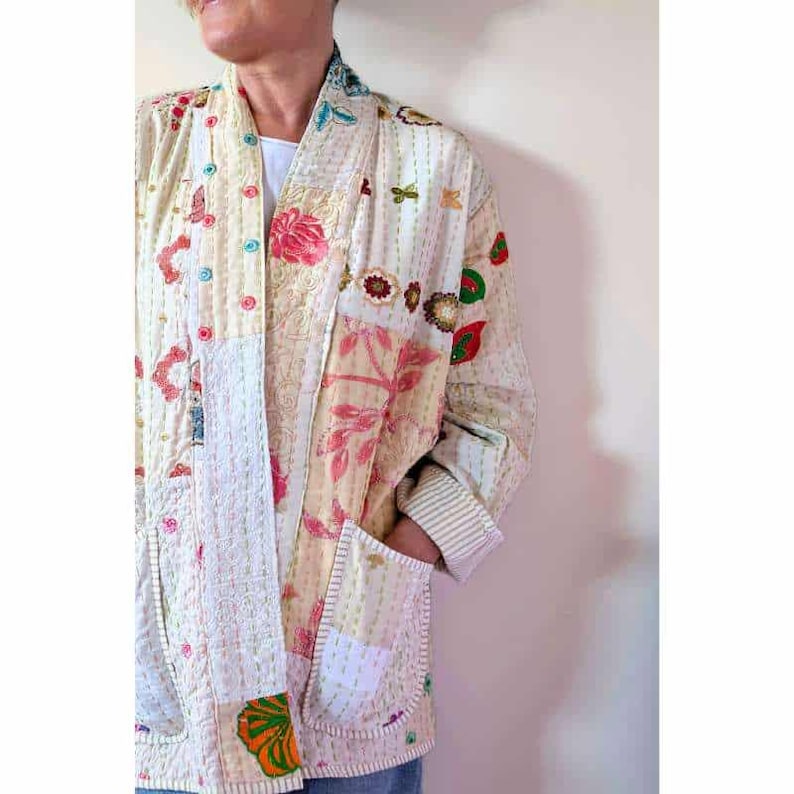 Puede incluir: Una chaqueta de manga larga con un dise&ntilde;o de patchwork en tonos crema, rosa y verde con motivos florales. La prenda incluye un cierre frontal con botones de colores, dos bolsillos delanteros y costuras blancas visibles en la tela.