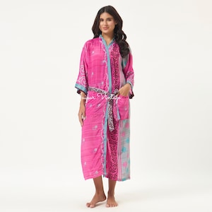 May include: A long-sleeved, calf-length robe in vibrant pink featuring a repeating white geometric pattern, a dark grey floral border along the front opening, and a matching tie belt. The garment includes a patch pocket and light blue trim.