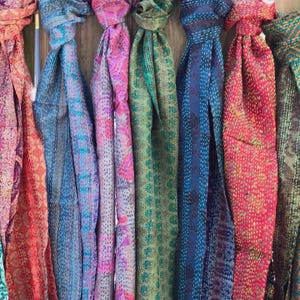 May include: A collection of seven long scarves featuring intricate kantha stitching and vibrant, multi-colored patterns. Each scarf displays a unique combination of floral, geometric, and abstract designs in shades of pink, blue, green, and orange.