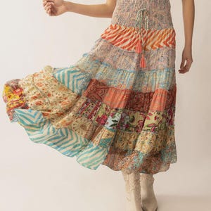 May include: A long, tiered skirt featuring a patchwork of various floral, paisley, and chevron patterns in shades of orange, blue, pink, and cream. The skirt has a high, smocked elastic waistband with a drawstring tie and a flared silhouette.