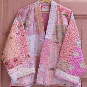 Peut inclure: Une veste matelassée présentant un motif patchwork dans diverses nuances de rose, pêche et blanc. Le vêtement est composé de multiples panneaux de tissu avec des détails de points de couture visibles, des manches longues et une ouverture frontale.