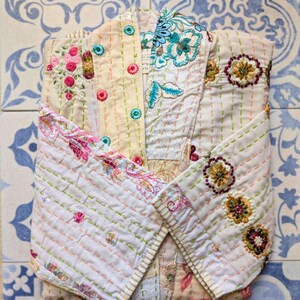 Puede incluir: Una chaqueta acolchada con base de color crema y bordados florales intrincados en tonos rosa, azul, verde y marr&oacute;n. La prenda presenta detalles de costura visibles, un cuello doblado y mangas largas con acentos bordados en toda la superficie.
