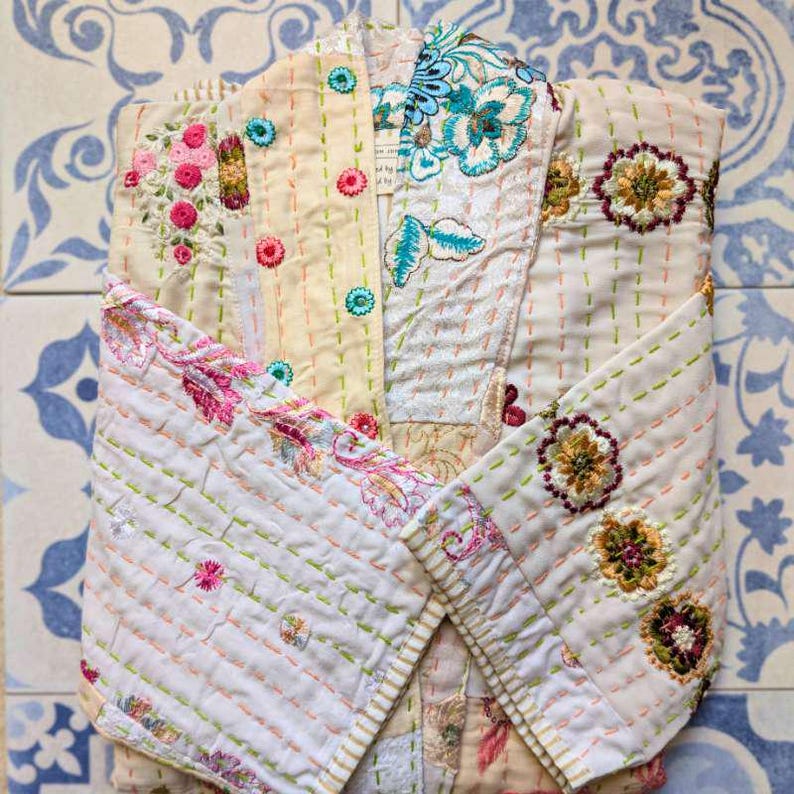 Puede incluir: Una chaqueta acolchada con base de color crema y bordados florales intrincados en tonos rosa, azul, verde y marr&oacute;n. La prenda presenta detalles de costura visibles, un cuello doblado y mangas largas con acentos bordados en toda la superficie.