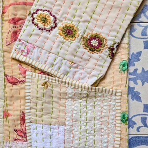 Puede incluir: Un textil acolchado con una base de color beige claro y l&iacute;neas de costura hechas a mano en naranja y verde. La tela muestra motivos florales bordados en tonos rosa, amarillo y verde, junto con parches de encaje blanco con perforaciones.