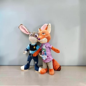 Pode incluir: Dois peluches representando um coelho e uma raposa. O coelho tem orelhas compridas, um uniforme de polícia azul com colete preto e calças azul-escuras. A raposa tem pelo laranja, uma camisa roxa de manga curta e calças bege claro.
