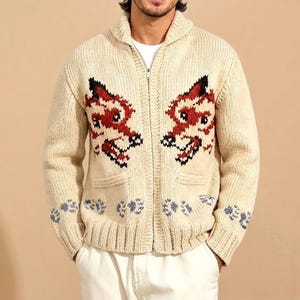 May include: A cream-colored, chunky knit zip-up cardigan featuring a shawl collar and long sleeves. The front displays two symmetrical, stylized fox head patterns in red, black, and white, with small paw print motifs along the bottom hem.