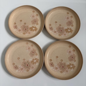 Op de afbeelding: Een set van vier ronde keramische borden in een lichtbeige gespikkelde afwerking. Elk bord heeft een bloemmotief in bruin- en tinten, gerangschikt in een groep aan de rechterkant. De borden hebben een gladde, licht verhoogde rand.