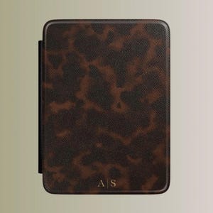 Puede incluir: Una funda rectangular para lector electrónico con un patrón de carey en marrón oscuro y negro con un acabado texturizado. En la parte inferior central de la funda se muestran las iniciales doradas A S separadas por una fina línea vertical.