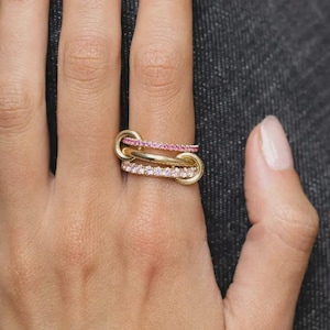 May include: A gold ring featuring three interconnected bands. The top band is set with small pink stones, the middle band is a plain polished gold loop, and the bottom band is set with small clear white stones, creating a layered, multi-textured appearance.
