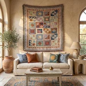 May include: A rectangular woven wall tapestry featuring a grid of squares with sun, moon, and wave motifs in shades of blue, orange, and cream, bordered by a Greek key pattern. The tapestry has a fringed edge and hangs above a beige sofa.