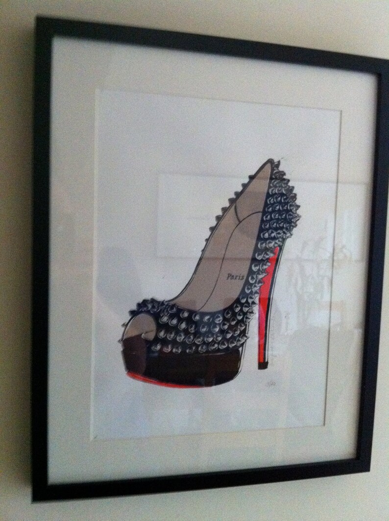 Puede incluir: Obra de arte enmarcada con una ilustraci&oacute;n de un zapato de tac&oacute;n alto. El zapato es negro con tachuelas plateadas, suela roja y tac&oacute;n rojo. La palabra "Par&iacute;s" est&aacute; impresa en el lateral. La obra est&aacute; en un marco negro.
