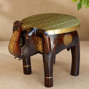 Puede incluir: Un taburete de madera marrón oscuro con forma de elefante y una tapa circular plana de latón con un patrón grabado. La figura del elefante tiene detalles de metal dorado en la trompa, las orejas y los costados, con cuatro patas firmes.