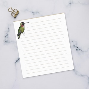 May include: A white rectangular notepad with horizontal lines and a small illustration of a green and red hummingbird in the top left corner. The notepad is positioned on a desk surface alongside a white keyboard, a coffee mug, and pencils.