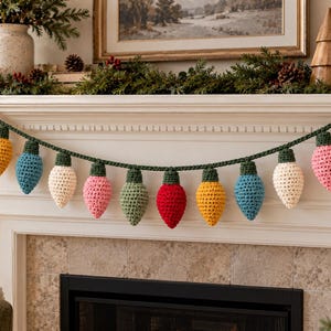 May include: A decorative garland featuring thirteen crocheted light bulb ornaments in various colours including red, yellow, blue, white, pink, and green, all attached to a dark green crocheted string with small dark green caps on each bulb.