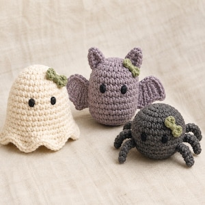 Pode incluir: Três figuras amigurumi de crochê: um fantasma cor de creme, um morcego lavanda com asas e uma aranha cinza-escura com oito pernas. Cada figura possui dois pequenos olhos de contas pretas e um pequeno laço de crochê verde-claro na cabeça.