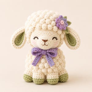 May include: A cream-coloured crocheted lamb plush toy featuring a textured bobble-stitch body, sage green inner ears and hooves, a lavender bow tied around its neck, and a small lavender flower accent attached to the side of its head.