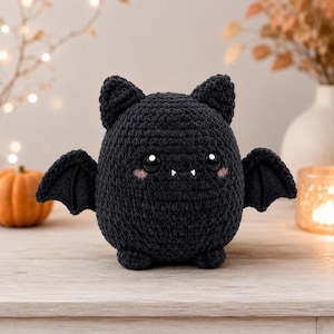 May include: A round, black crochet bat plush toy featuring two pointed ears, small wings, and two small feet. The bat has large, glossy black eyes, two small white fangs, and soft pink blush marks on its cheeks, all crafted with a textured yarn finish.