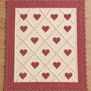 May include: A rectangular crochet blanket featuring a cream-colored center with a diamond grid pattern formed by raised cream bobbles. Each diamond contains a textured dark rose-colored heart. The blanket has a wide dark rose scalloped border.