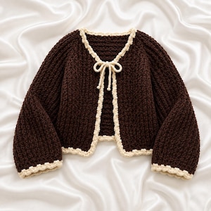May include: A dark brown crochet cardigan featuring a textured knit pattern, long sleeves, and a cream-colored scalloped trim along the edges. The front opening is secured with a matching cream-colored crochet tie closure at the neckline.
