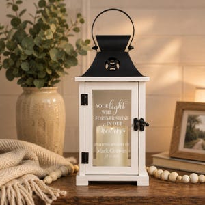 May include: A white decorative lantern with a black metal top and handle. The glass pane features white text that reads: Your light will forever shine in our hearts, In loving memory of Mark Corwin, 1970-2026. A cream-colored pillar candle sits inside.
