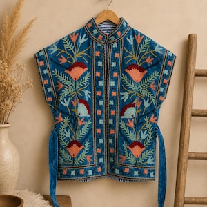 Op de afbeelding: Een mouwloos blauw vest met een mandarijnkraag en ingewikkeld bloemenborduursel in tinten rood, oranje en lichtblauw. Het kledingstuk heeft een opening aan de voorzijde en lange bijpassende blauwe stoffen linten aan de zijkanten, met een blauw-wit gestreepte voering.