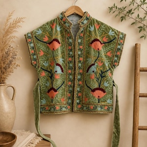 May include: A sleeveless olive green velvet waistcoat featuring intricate floral embroidery in shades of coral, light blue, and dark navy. The garment has a mandarin collar, a front opening, and long matching velvet tie straps at the side seams.