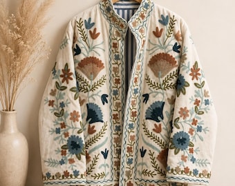 Puede incluir: Una chaqueta acolchada de color crema con intrincados bordados florales en tonos azul, óxido y verde. La chaqueta tiene cuello mao, mangas largas y una abertura frontal que revela un forro interior con rayas azules y blancas.