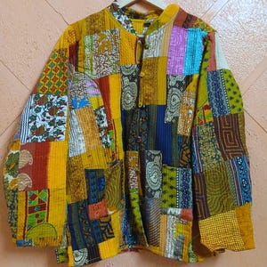 Op de afbeelding: Een doorgestikt jack met lange mouwen en een levendig patchworkontwerp met diverse kleurrijke stoffen vierkanten in tinten geel, blauw, groen en paars. Het jack heeft een mandarijnkraag, een knoopsluiting aan de voorzijde en twee zakken.