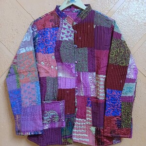 Op de afbeelding: Een doorgestikt jack met een patchworkontwerp van diverse stofpatronen in tinten paars, magenta, bordeauxrood en blauw. Het jack heeft een opstaande kraag, lange mouwen, een knoopsluiting aan de voorzijde en twee opgestikte zakken voorop.