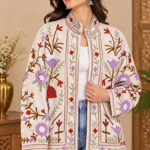 May include: An off-white jacket featuring a mandarin collar and long sleeves, decorated with vibrant floral embroidery in shades of purple, red, and green. The open-front design showcases intricate botanical patterns along the edges and sleeves.