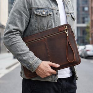 May include: A dark brown leather document case featuring a horizontal zippered front pocket, a matching leather wrist strap attached with a gold-toned metal clasp, and visible stitching along the center and edges of the rectangular bag.