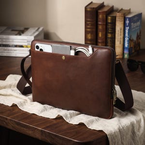May include: A brown leather messenger bag with a zippered main compartment and an external front pocket secured by a brass snap. The front pocket contains a white smartphone, a silver power bank, and a white charging cable with a USB-C connector.