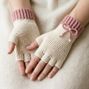 May include: A pair of fingerless gloves crafted from cream-colored yarn with a ribbed cuff in a dusty rose shade. The gloves feature a small crocheted bow detail on the back of the hand, accented with two small white faux pearls at the ends.