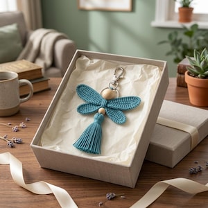 May include: A teal dragonfly-shaped macrame keychain featuring crocheted wings, two smooth wooden beads, and a matching tassel, attached to a silver-toned metal lobster clasp, presented inside a light gray gift box with white tissue paper.