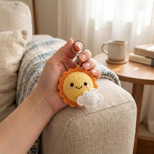 May include: A crocheted sun keychain featuring a bright yellow circular face with black bead eyes, a stitched smile, and pink cheeks. The sun has an orange scalloped border and a small white crocheted cloud attached to the lower right side.