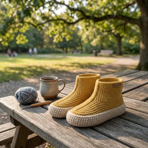 Puede incluir: Un par de botines de ganchillo color amarillo mostaza con suelas gruesas de color crema. Los botines presentan un patrón de punto texturizado y una pequeña etiqueta rectangular marrón en el lateral. Junto a ellos hay una taza y lana.