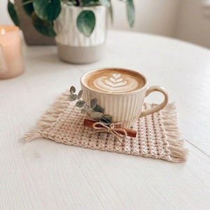 May include: A cream-colored, vertically ribbed ceramic mug filled with latte art, resting on a square, woven beige coaster with fringed edges. A small cinnamon stick and a sprig of dried eucalyptus are tied to the mug with thin twine.