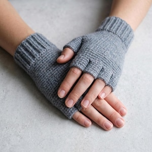 May include: A pair of light gray fingerless gloves featuring a textured knit pattern and a ribbed cuff design. The gloves cover the back of the hand and the wrist, leaving the fingers and thumb exposed for dexterity while providing warmth to the hands.