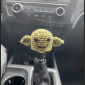 May include: A light green crocheted cover for a car gear shift knob, featuring a rounded head shape, two pointed ears, and embroidered black thread details forming two small eyes and a curved mouth, positioned atop a black leather gear shifter.