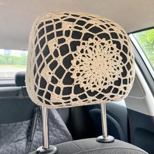 May include: A beige crochet headrest cover featuring an intricate circular mandala pattern in the center, surrounded by a geometric grid design. The cover is fitted over a black car headrest, which is supported by two polished metal posts.