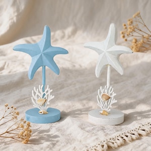 Op de afbeelding: Twee decoratieve zeesterornamenten op standaards. De ene zeester is lichtblauw en de andere is wit. Elke zeester is gemonteerd op een dunne staaf boven een witte koraalvormige basis met kleine bevestigde schelpen, op een ronde houten voet.
