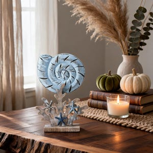 May include: A decorative tabletop ornament featuring a light blue, distressed-finish nautilus shell mounted on a base shaped like white and brown coral branches, accented with three small, dark blue starfish attached to the coral structure.