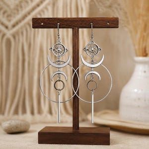 May include: A pair of silver-toned dangle earrings featuring a vertical arrangement of a stylized eye symbol, a crescent moon, a circular hoop, and a small ring ending in a straight bar. The earrings are suspended from hooks and display a metallic finish.