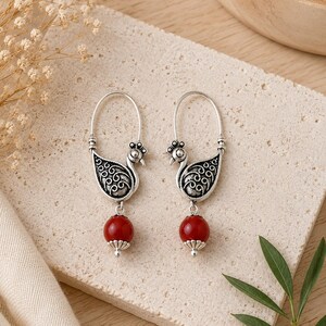 May include: A pair of silver-toned hoop earrings featuring a stylized bird motif with intricate scrollwork patterns on the body. Each earring is accented with a single, smooth, round red bead suspended from the base of the bird-shaped metal frame.
