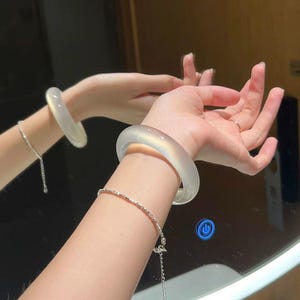 Puede incluir: Una pulsera rígida de color blanco translúcido y superficie lisa con perfil redondeado, acompañada de una delicada pulsera de cadena plateada con pequeños eslabones metálicos reflectantes. La pulsera tiene un acabado pulido y color blanco lechoso.