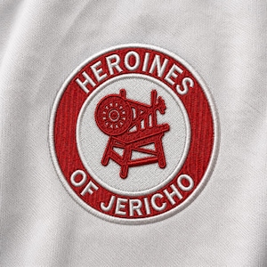 May include: A circular embroidered patch on a white fabric background. The patch features a red border with the white text HEROINES OF JERICHO and a central red graphic of a spinning wheel. The design is stitched with a visible thread texture.