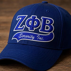 Op de afbeelding: Een koningsblauwe baseballpet met de Griekse letters ZΦB geborduurd in blauw met een witte omlijning. Onder de letters bevindt zich een gebogen blauwe banner met de witte geborduurde tekst Sorority Inc. in een schrijflettertype met witte rand.