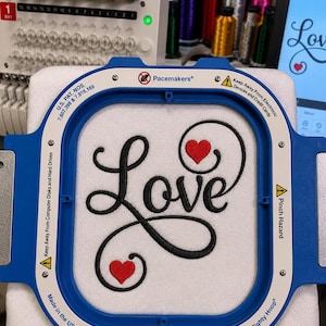 May include: A blue Mighty Hoop embroidery frame holding white fabric with the black embroidered word Love and two small red hearts. The frame features safety warnings, patent numbers, and the text Made in the USA with a small flag icon.
