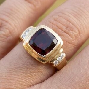 Op de afbeelding: Een gouden ring met een grote, kussengeslepen, dieprode granaat edelsteen in een gladde zetting. De ringband is aan weerszijden van de centrale steen bezet met drie kleine, ronde, helderwitte diamanten voor een gebalanceerd ontwerp.