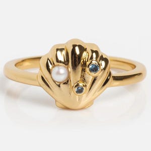 Pode incluir: Um anel dourado com um design central em forma de concha. A concha é adornada com uma pérola branca redonda no lado esquerdo e duas pequenas pedras preciosas azuis redondas posicionadas verticalmente no lado direito da superfície da concha.